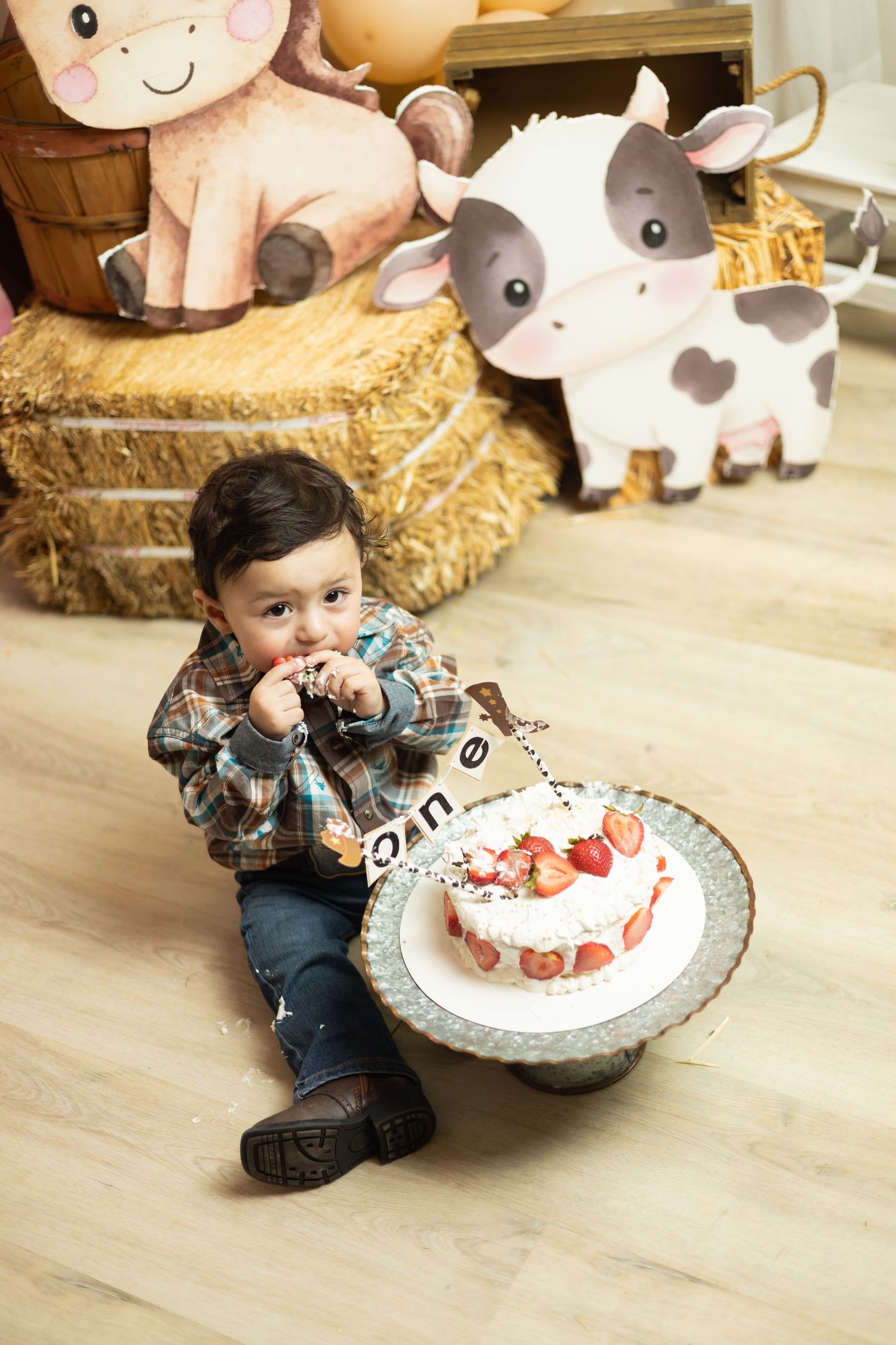 Toddler boy eating strawberry birthday cake at farm-themed first birthday party with hay bales and cow decorations.