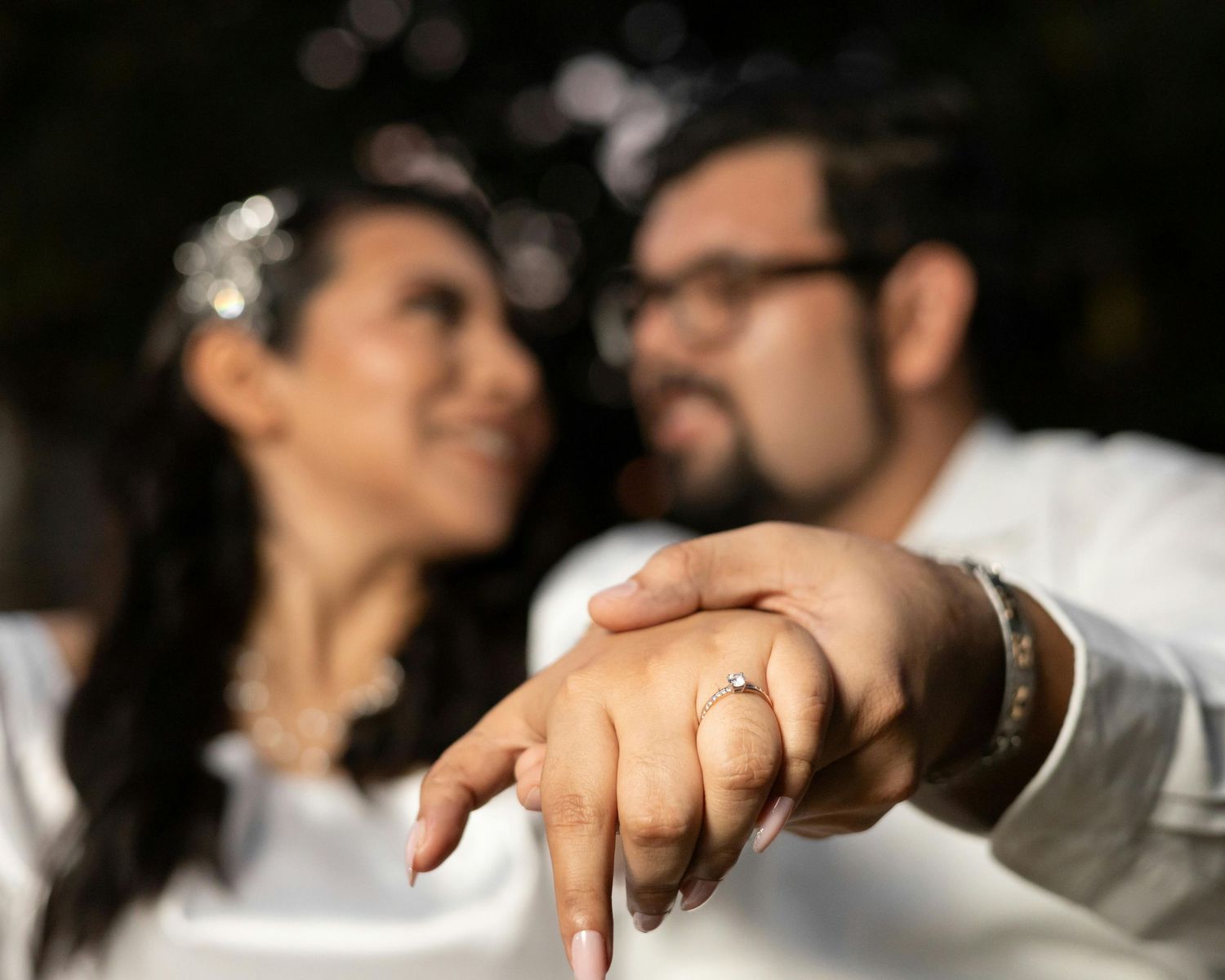 Couple in white outfits holding hands showing engagement ring, smiling outdoors in romantic blurred background.