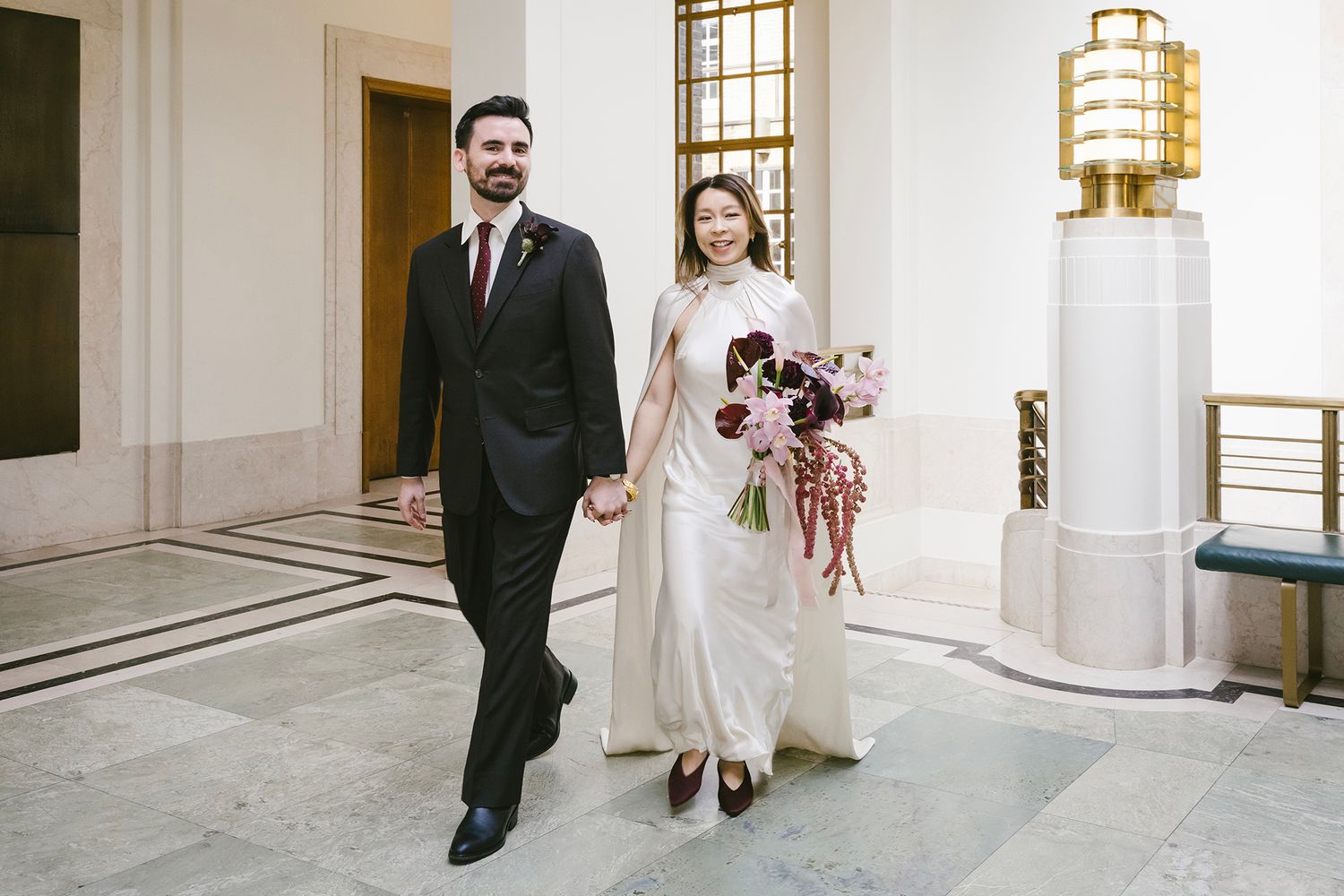 Bride in white gown holding floral bouquet walks hand-in-hand with groom in black suit at elegant Hackney Town Hall wedding venue.