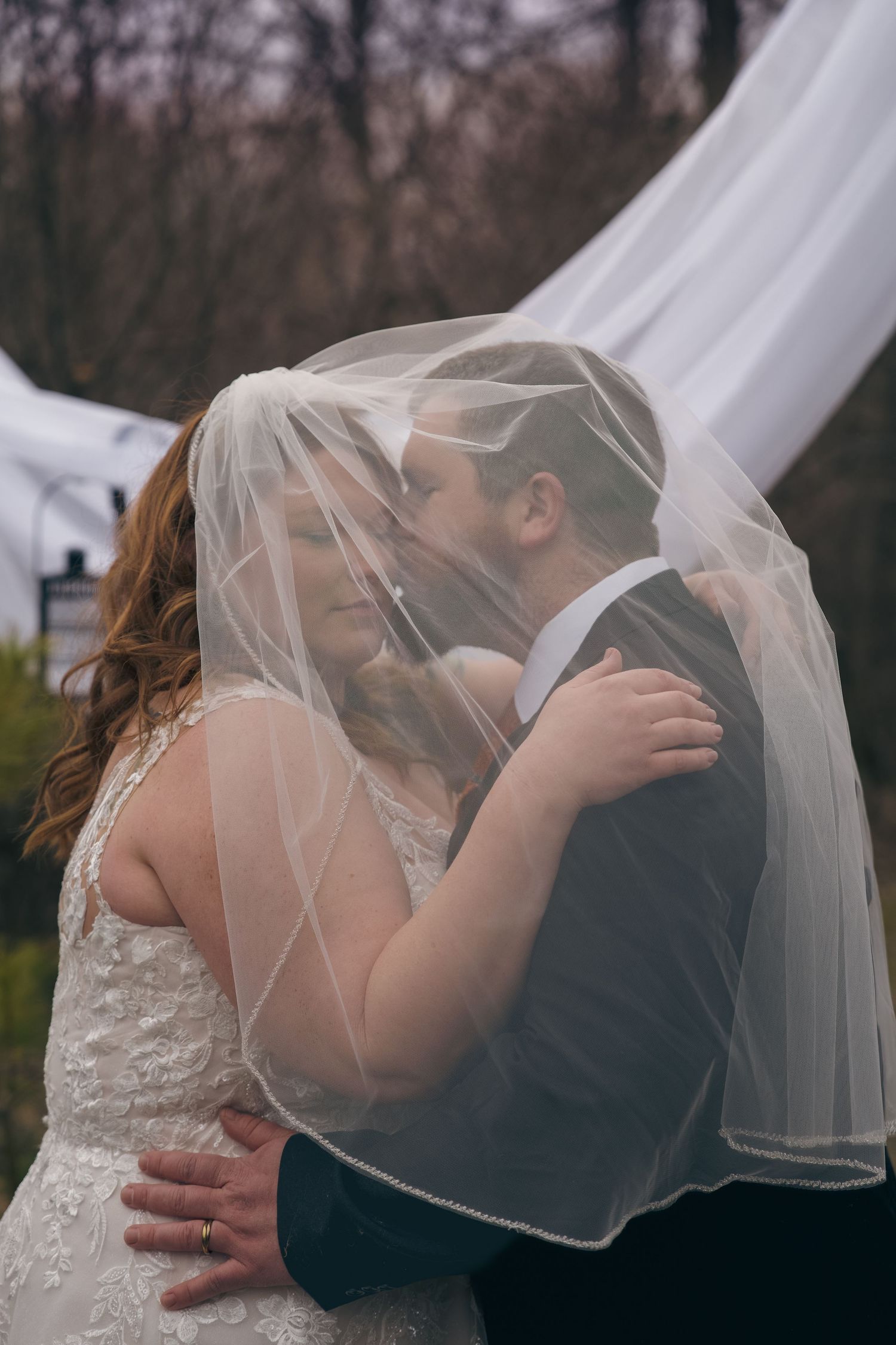 A bride and groom share a romantic kiss under a flowing white veil outdoors on their wedding day.