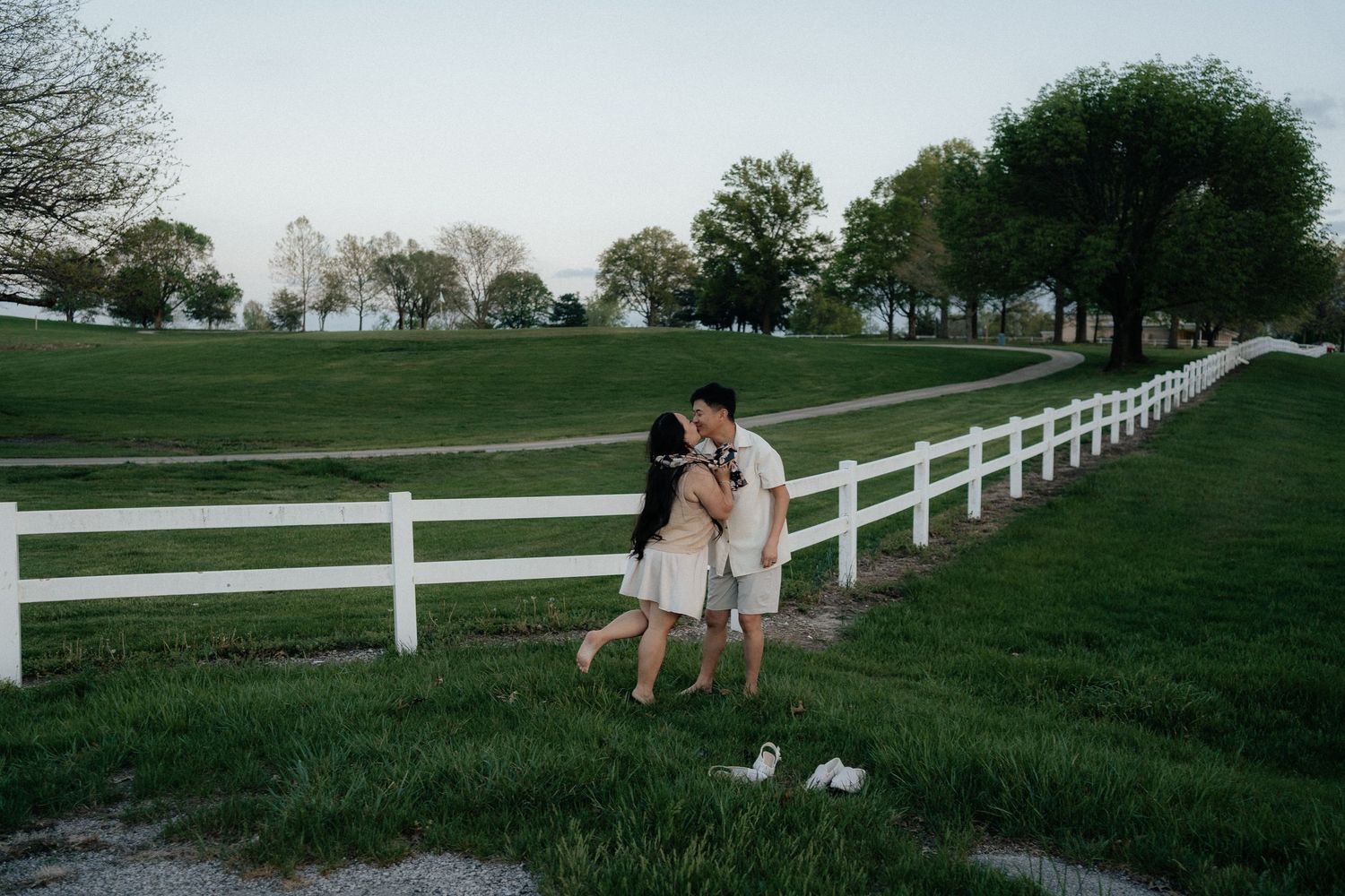 Couple in white outfits holding hands and dancing barefoot on green grass beside a white fence at dusk.