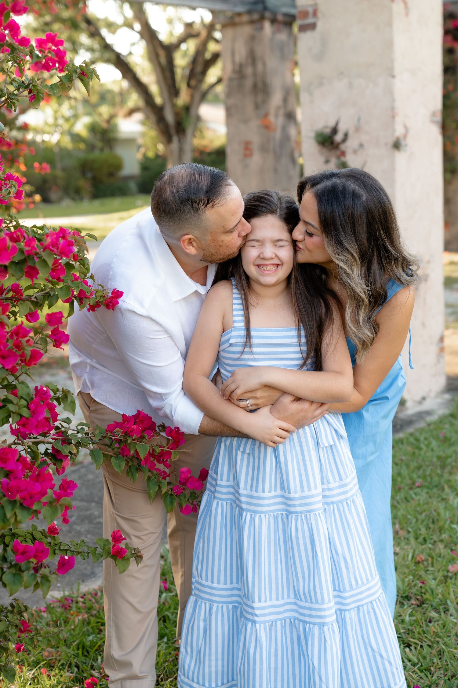 Parents kissing their smiling daughter on cheeks outdoors near pink bougainvillea flowers in a garden setting.