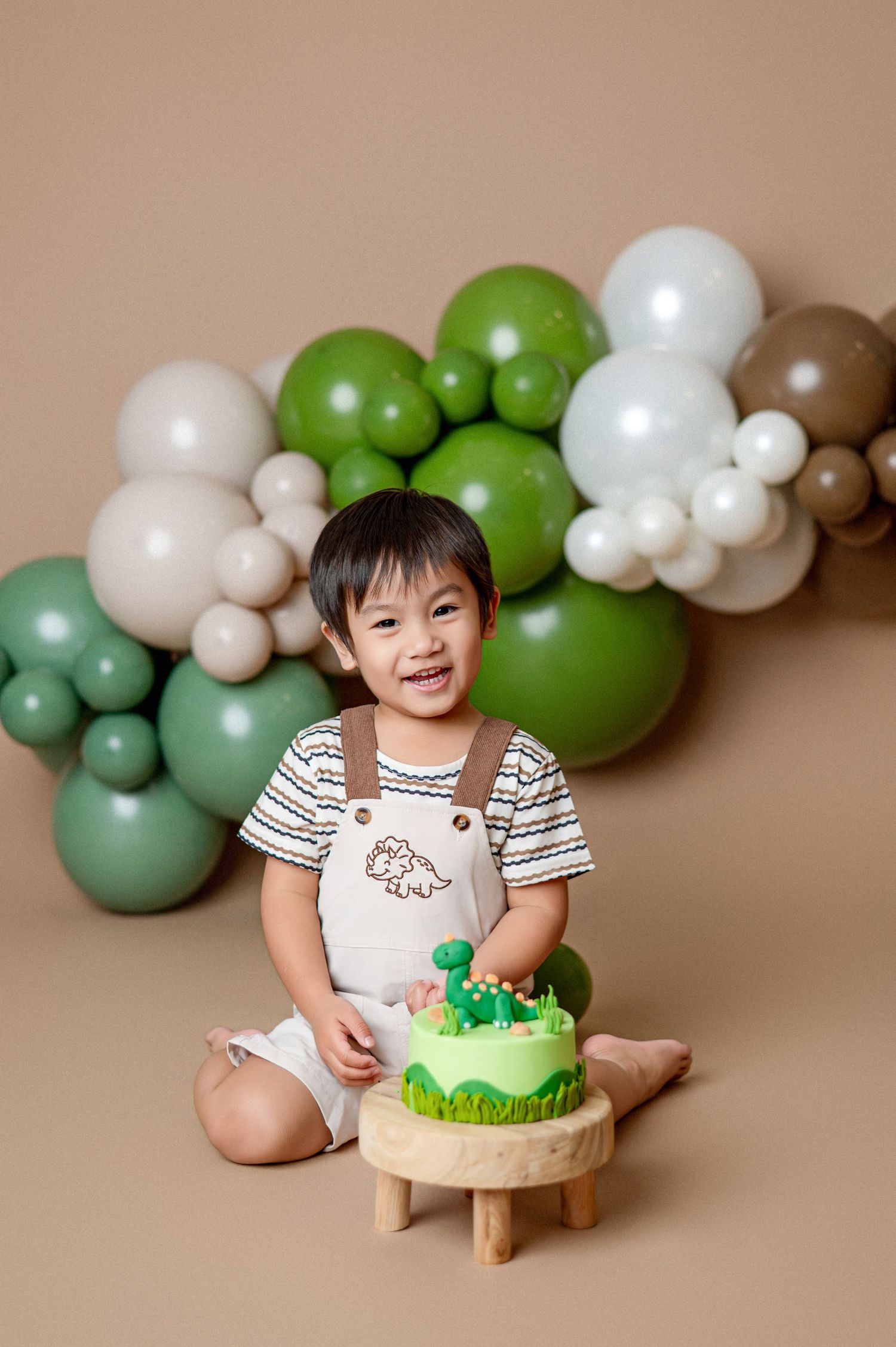 Baby girl in pink dress celebrates first birthday with cake and colorful balloon garland backdrop.
