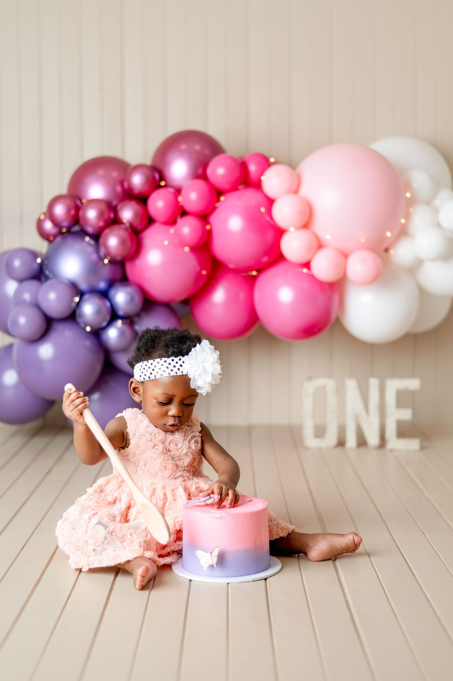 Baby girl in pink dress celebrates first birthday with cake and colorful balloon garland backdrop.
