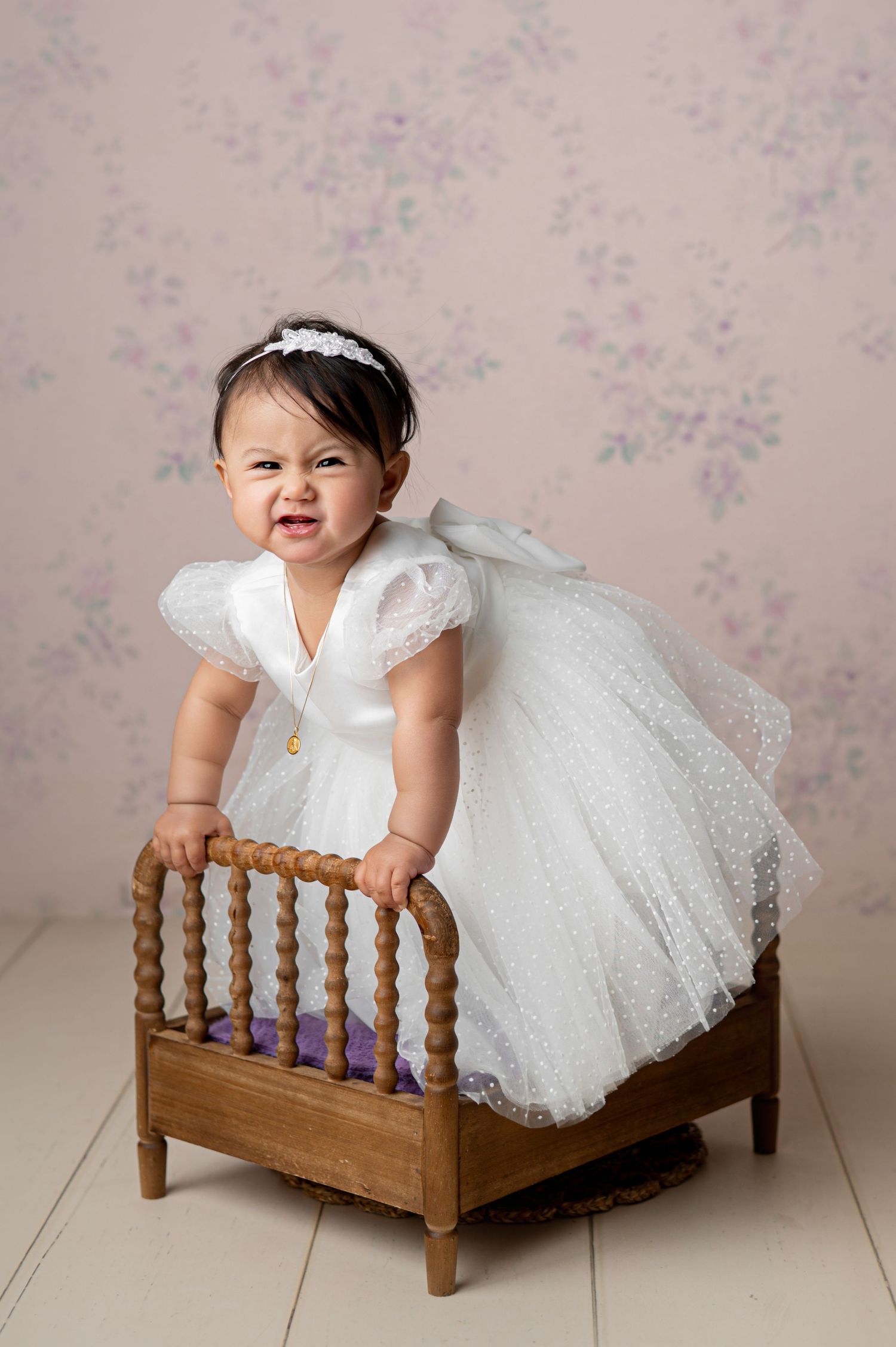 Smiling baby girl in floral romper and bow headband crawling on white wooden floor near sheer curtains.