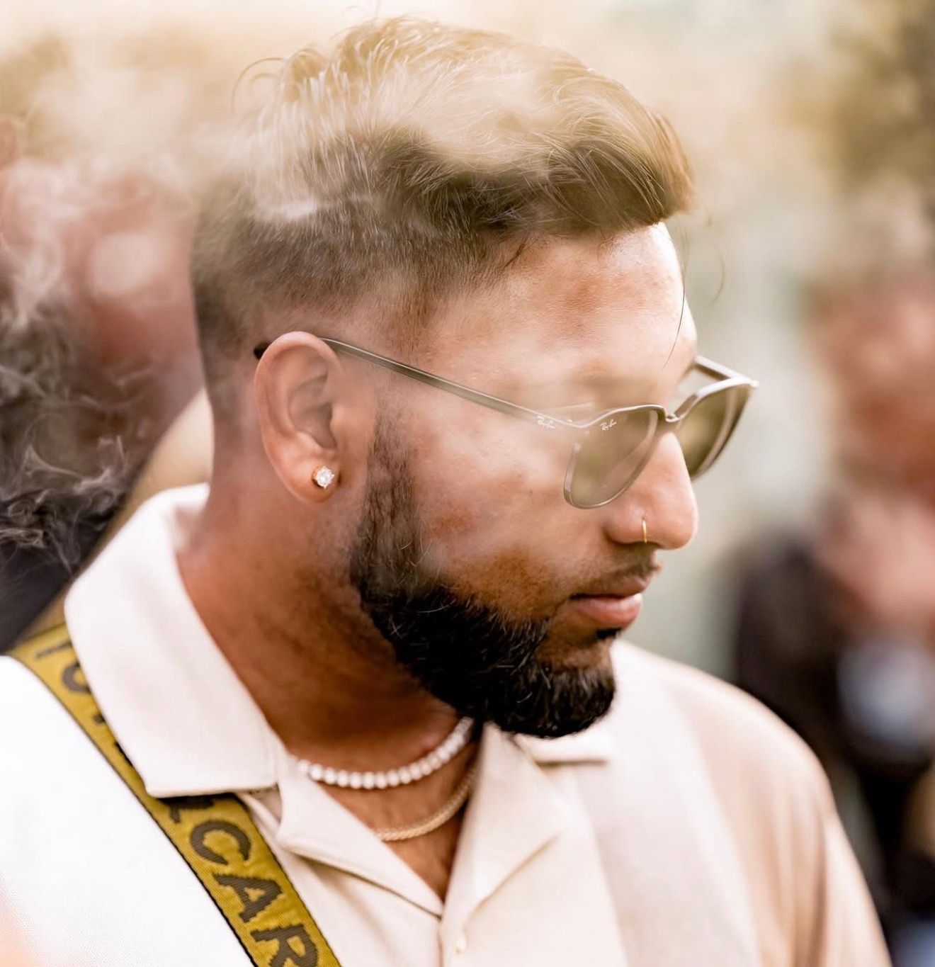 Stylish bearded man wearing sunglasses and pearl necklace at an outdoor event.