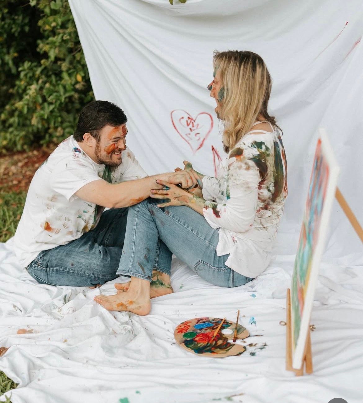 Couple sitting on white sheet outdoors, covered in paint, laughing and holding hands near a heart drawing.
