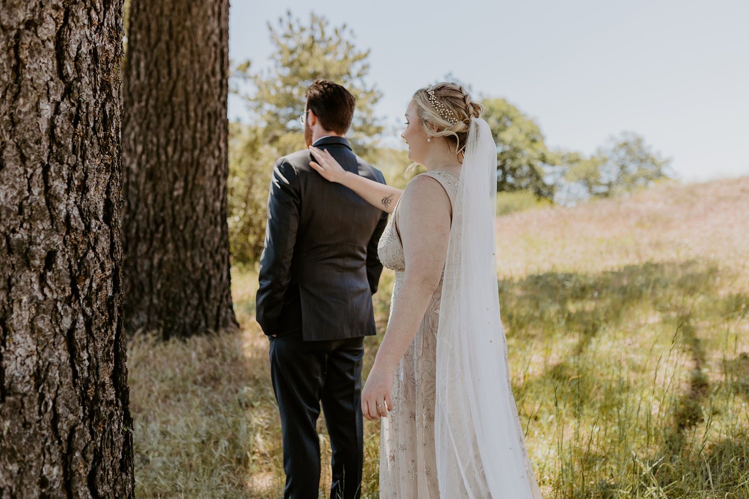 Bride and groom stand together near a tree in an outdoor meadow, bride wearing a flowing white gown and veil at Lake Cuyamaca, Julian.