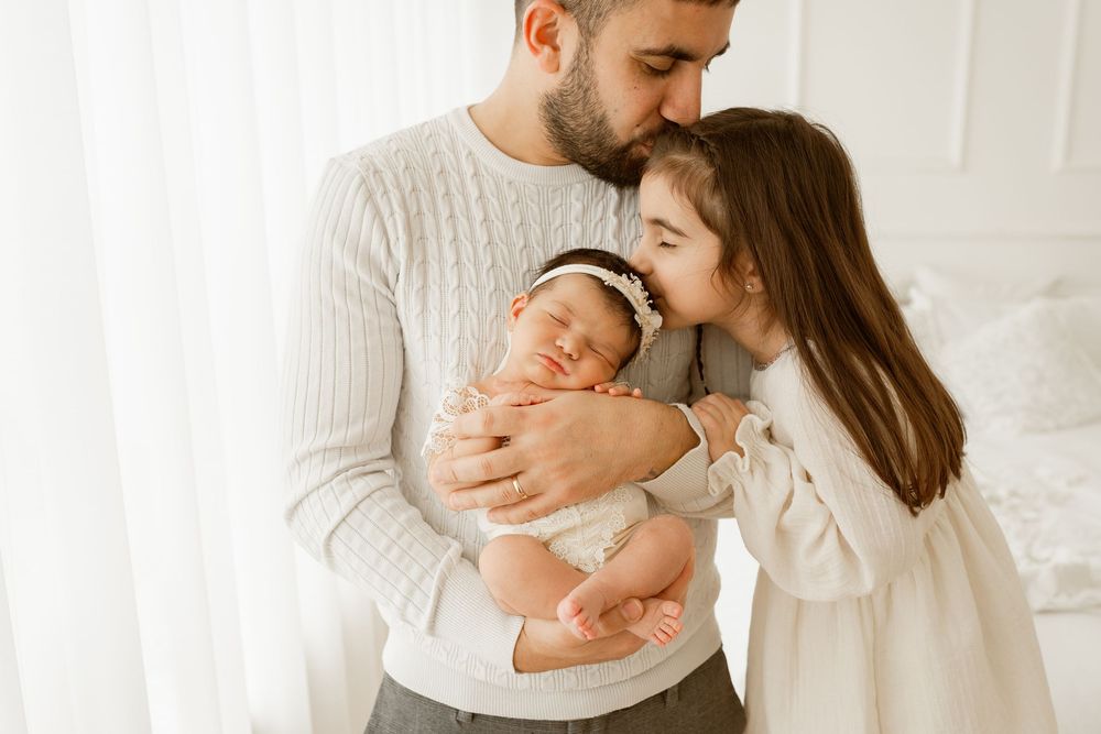 A father holds a sleeping newborn baby while an older sibling kisses the infant in a bright, white room.