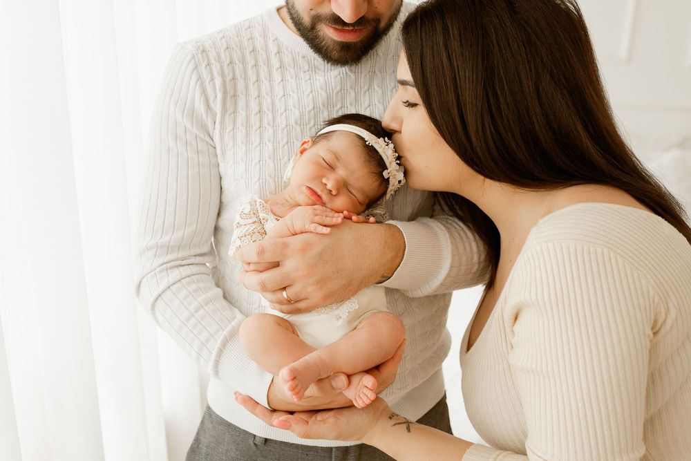 Parents tenderly holding and kissing their sleeping newborn baby wrapped in white.