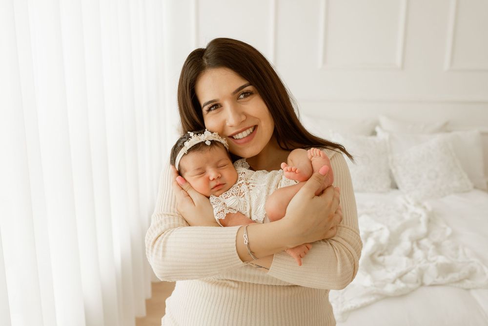 Smiling brunette mother holding sleeping newborn baby in white headband in a bright white bedroom.