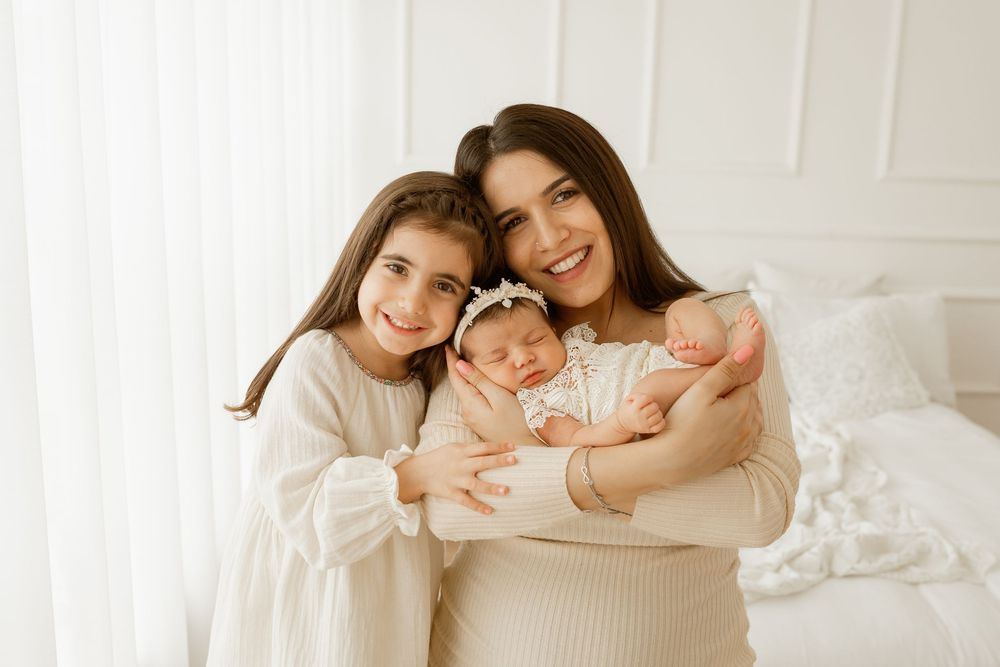 A smiling mother holds twin newborns while her young daughter embraces them in a bright white room.