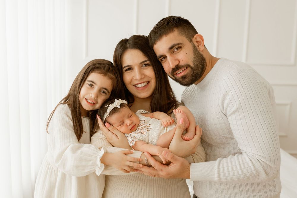 Family of four in white outfits poses together, parents holding newborn baby girl with floral headband and older daughter smiling.