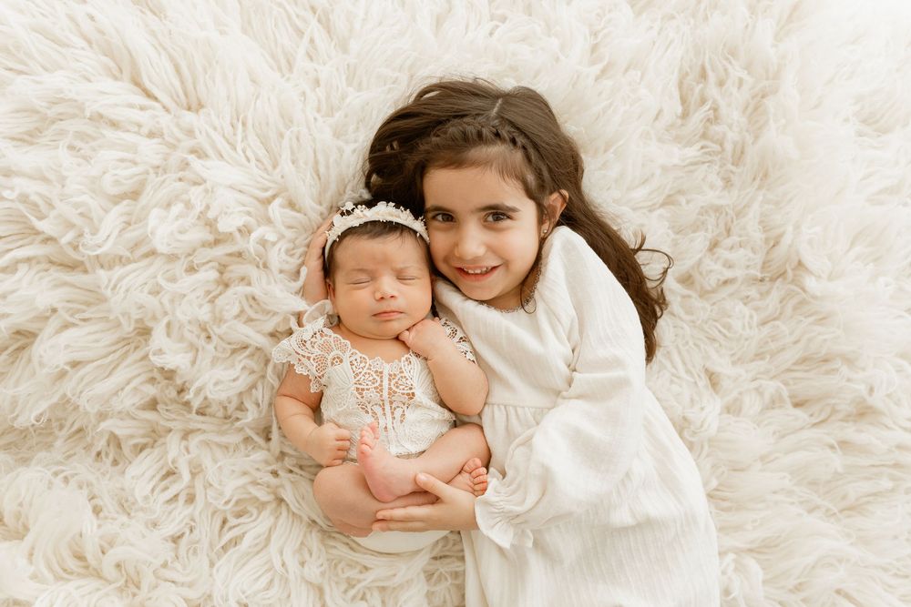 Older sister in white dress lovingly holds sleeping newborn on fluffy cream rug in professional photo.