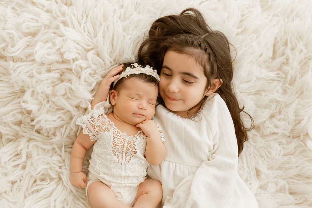 Older sister in white dress cuddles sleeping newborn baby girl in lace romper on fluffy cream rug.