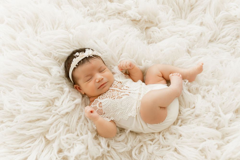 Peaceful newborn baby girl sleeping on fluffy white rug wearing lace romper and floral headband.