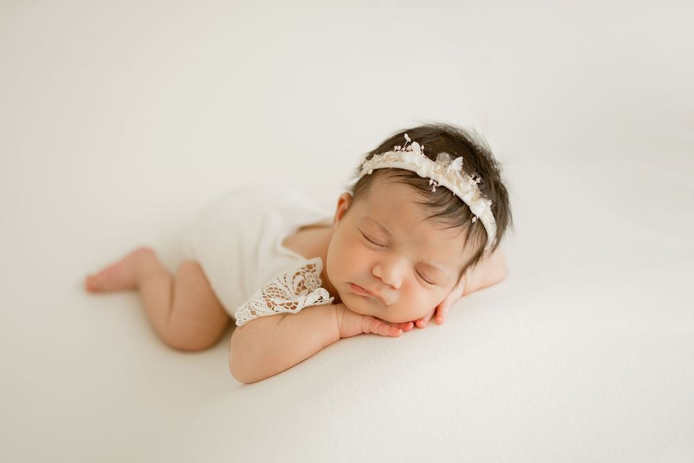 Sleeping newborn baby girl in white lace romper and floral headband posed on white backdrop.