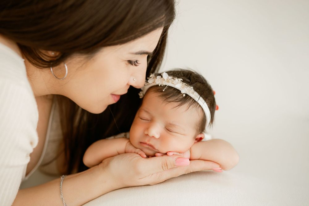 A mother tenderly gazes at her sleeping newborn baby wearing a floral headband in a soft white studio setting.