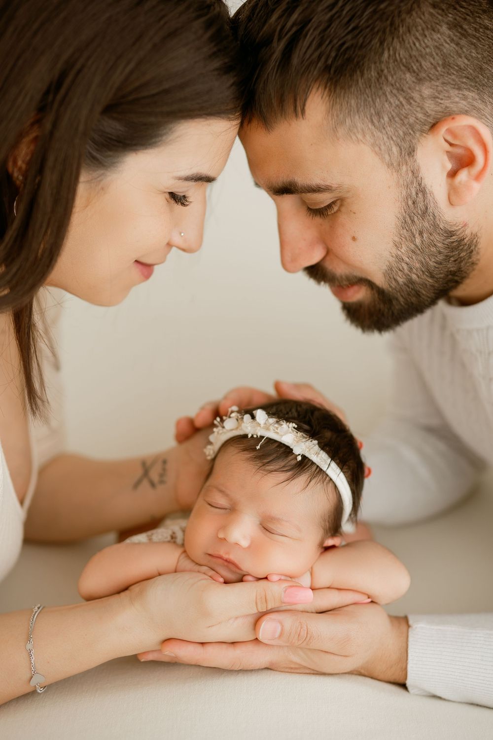 Parents tenderly lean foreheads together over their sleeping newborn baby girl wearing a delicate floral headband.