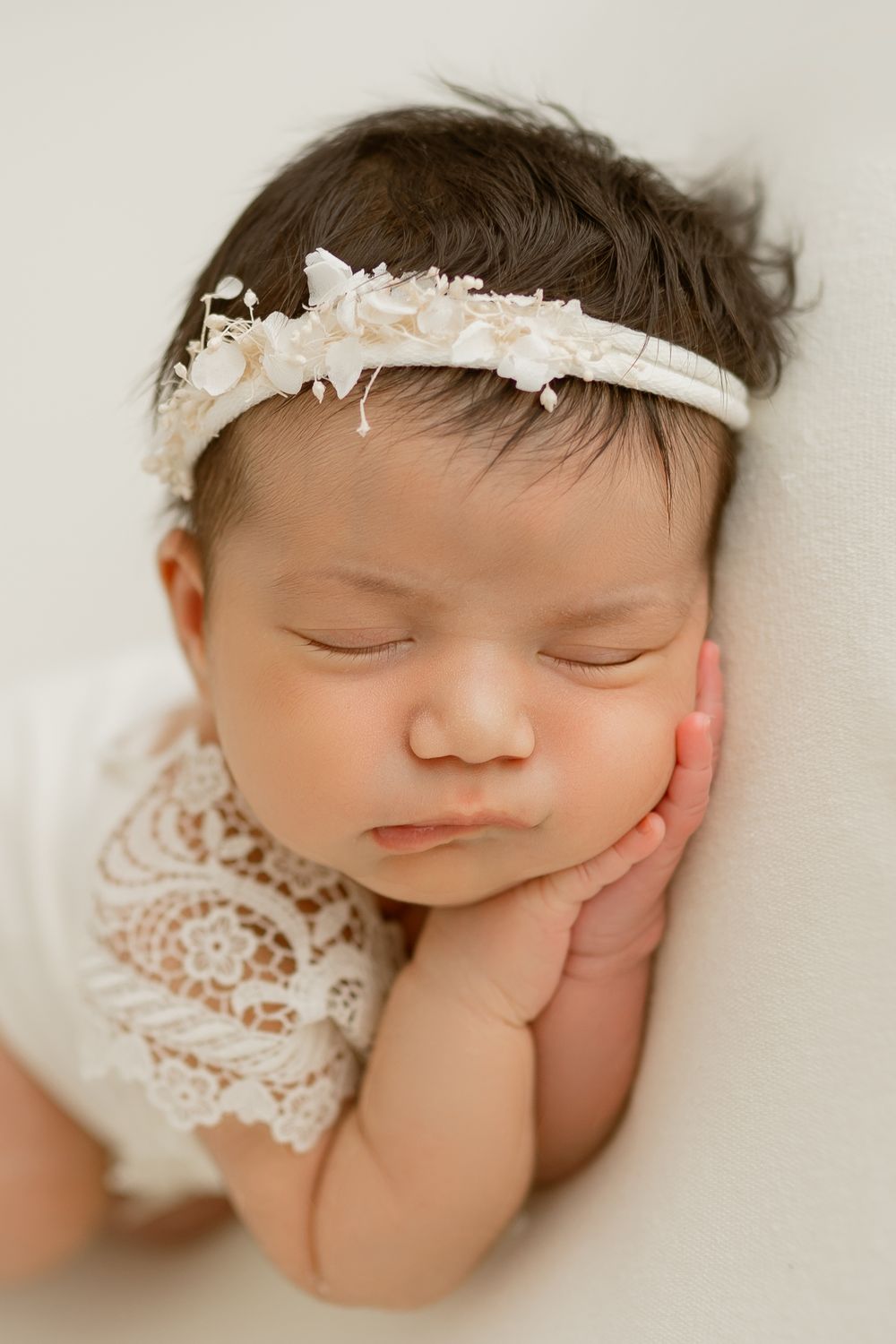 Sleeping newborn baby girl wearing white lace romper and floral headband, resting cheek on hands against soft white background.
