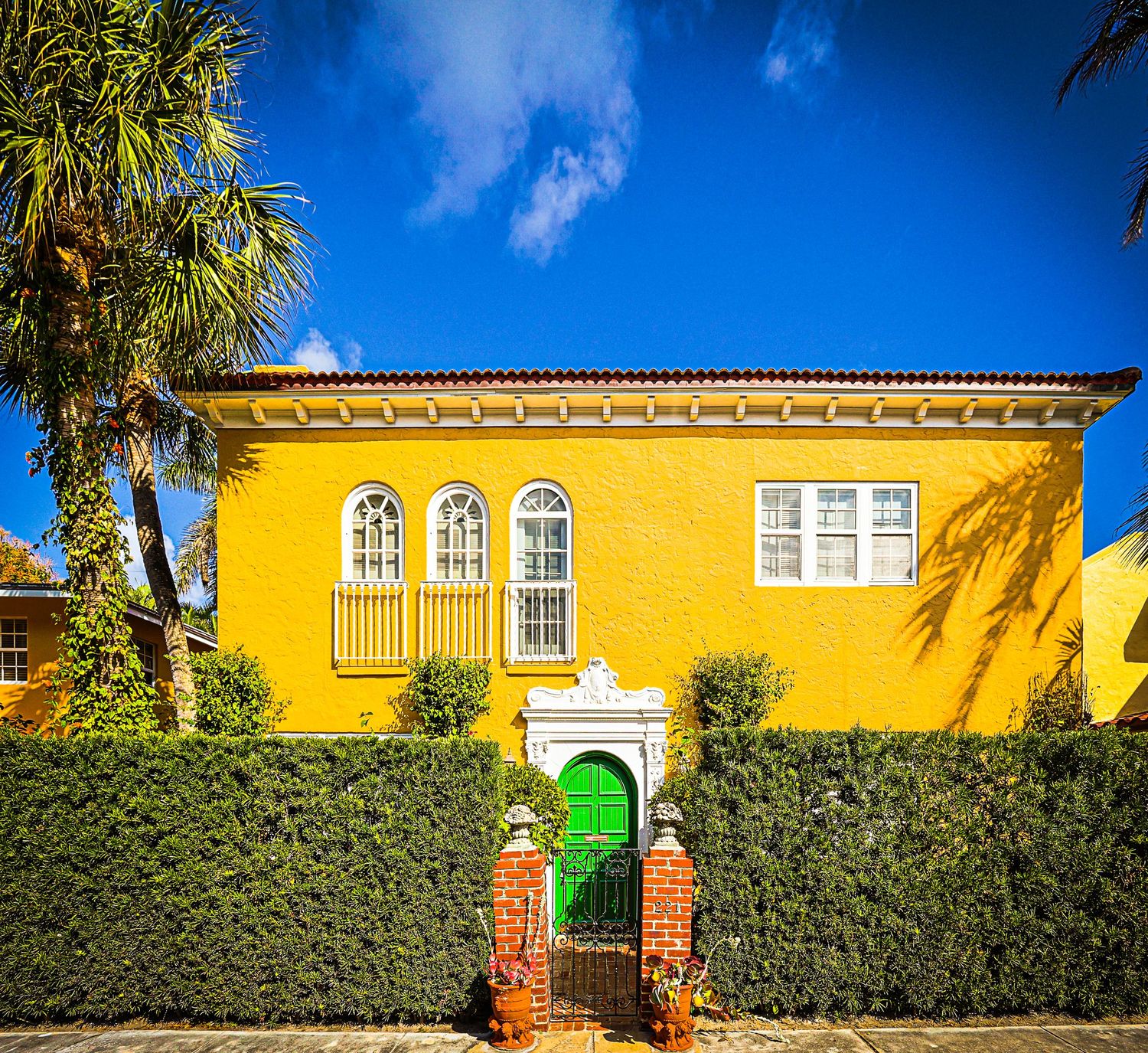 Vibrant yellow Mediterranean-style home with green gate, arched windows, ivy hedges, and palm trees under a bright blue sky.