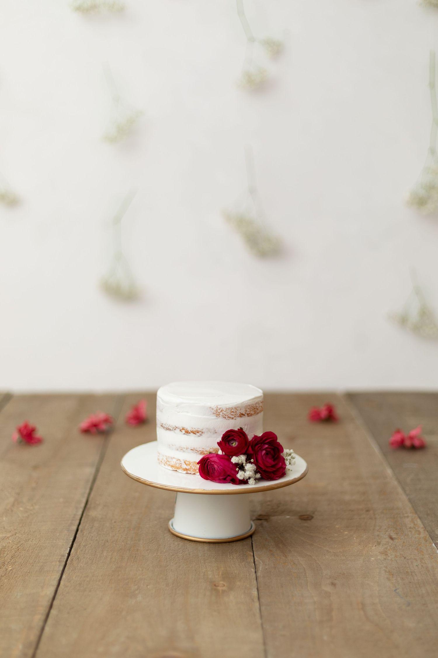Simple naked cake decorated with fresh red flowers on a white cake stand against wooden backdrop.