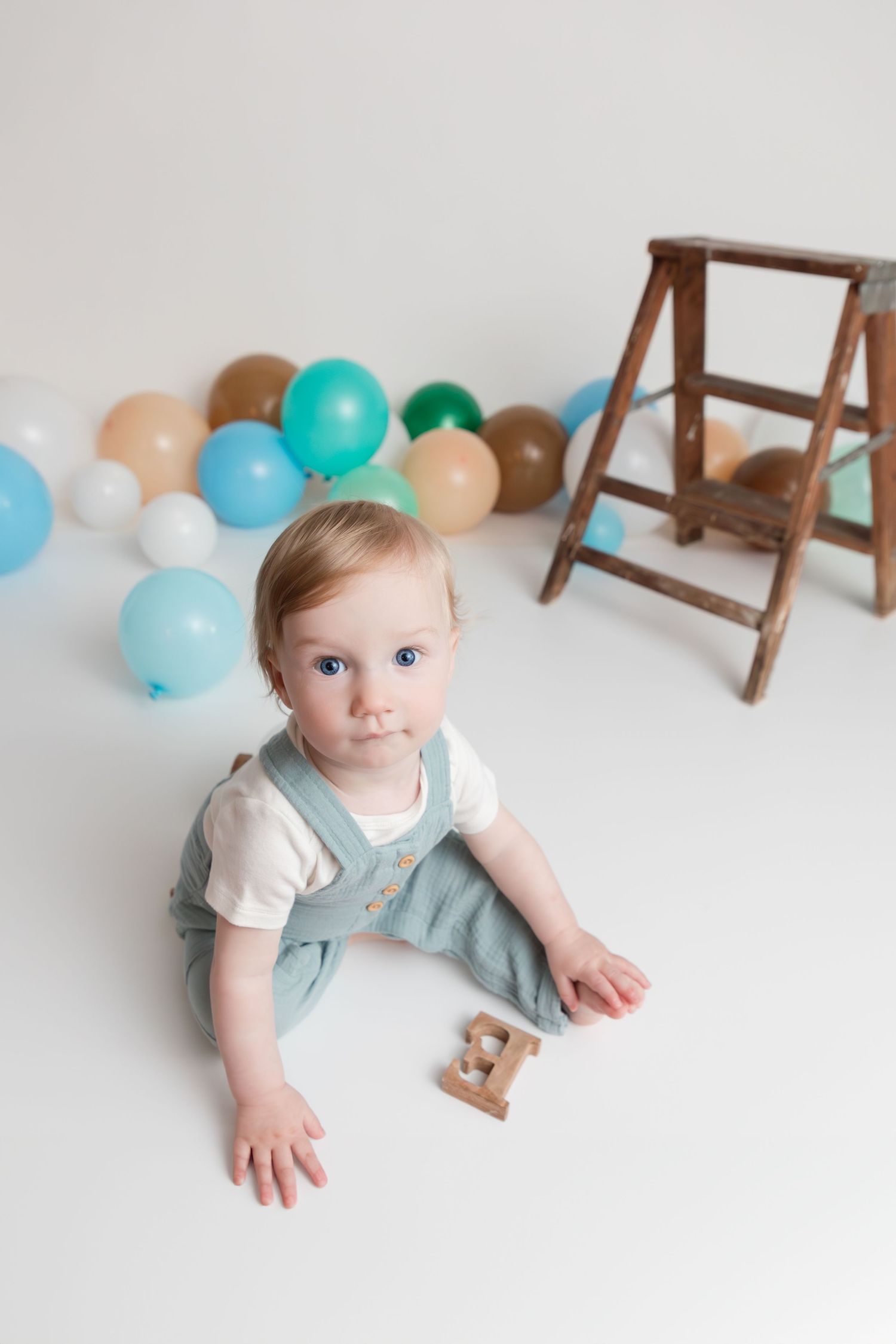 First birthday setup with neutral colored balloons, wooden ladder, and cake display on white background.