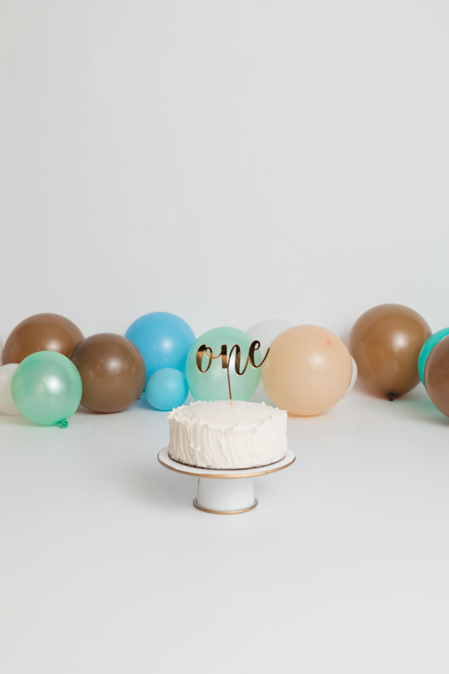 White birthday cake with 'one' topper surrounded by brown, blue, and mint colored balloons on neutral background.