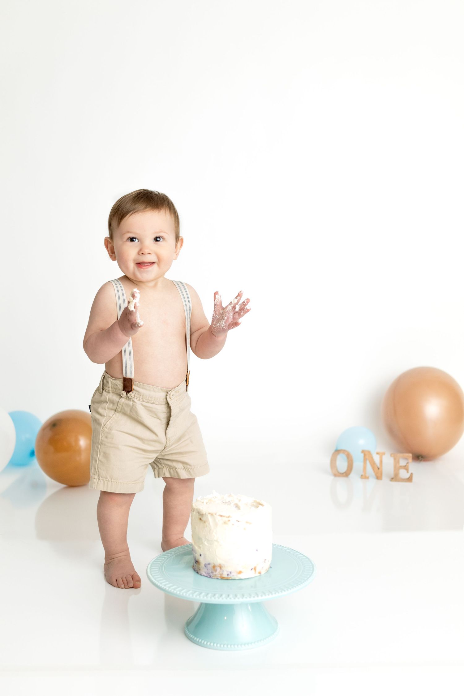 Minimalist cake smash photo setup with beige suspender shorts and blue cake stand.