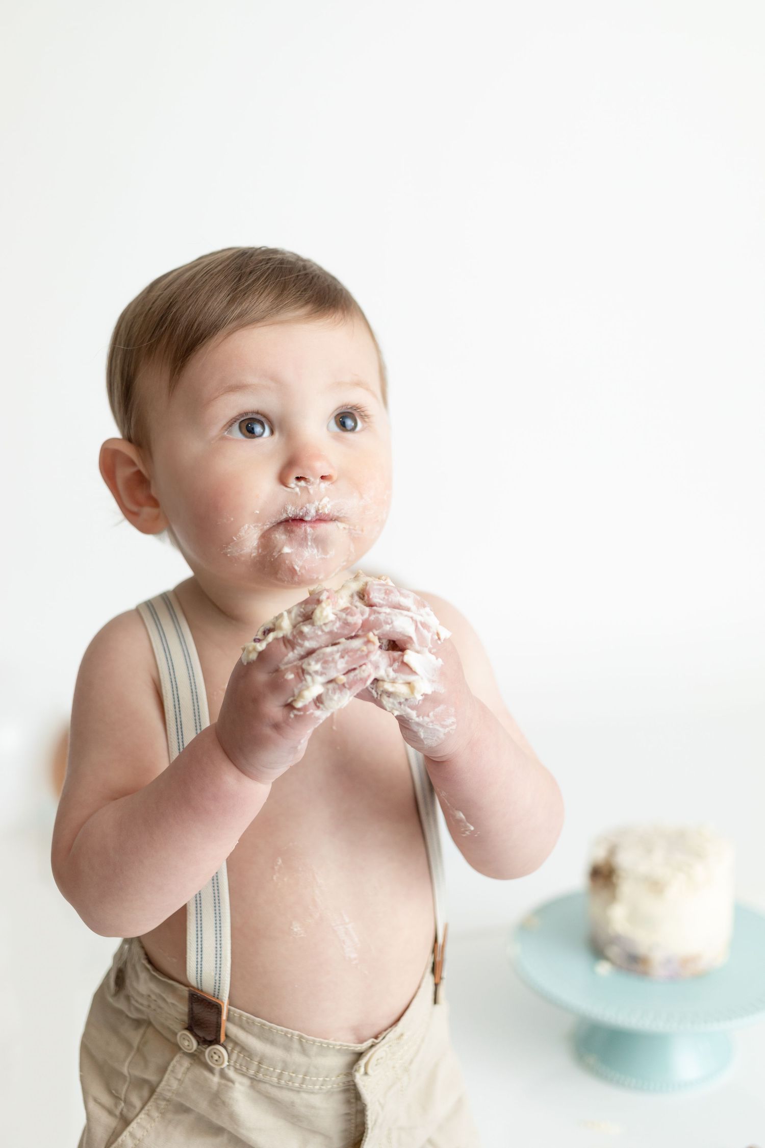 Clean white cake smash photo setup with vintage suspenders and turquoise cake stand.