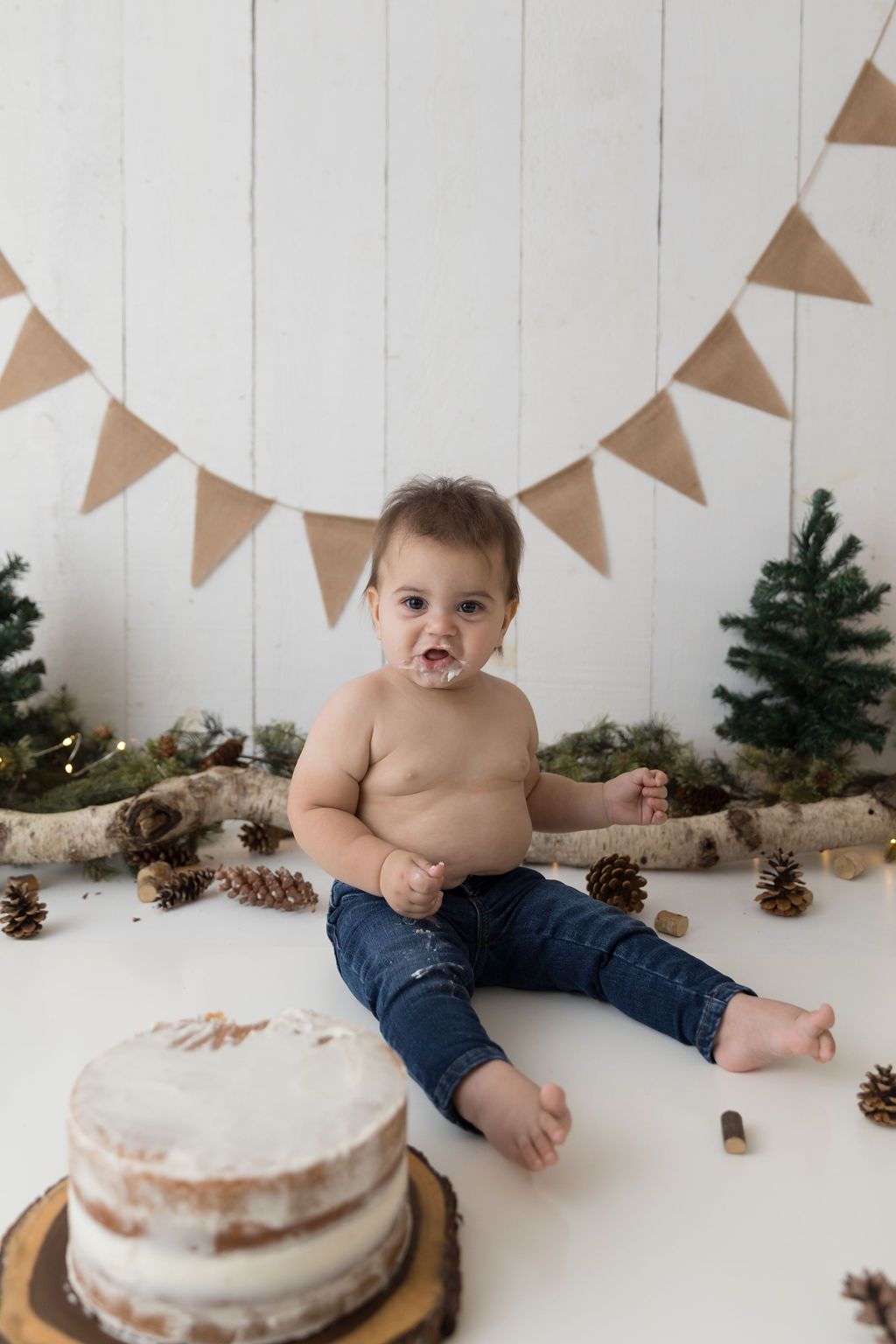A cake smash birthday photo setup with pinecones, evergreen trees and burlap banner decorations.