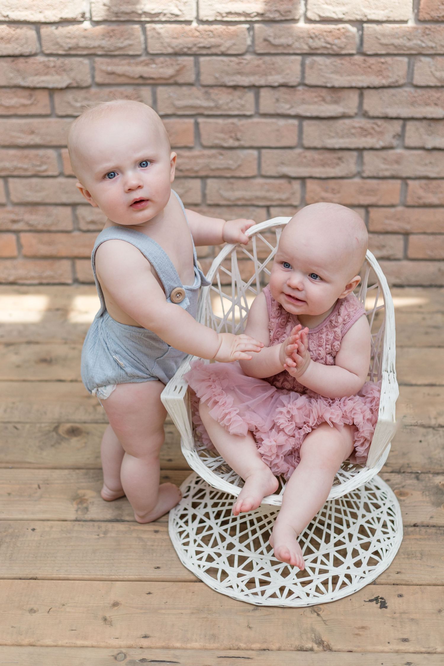 Two babies in summer outfits pose together on a white wicker chair against a brick wall background.