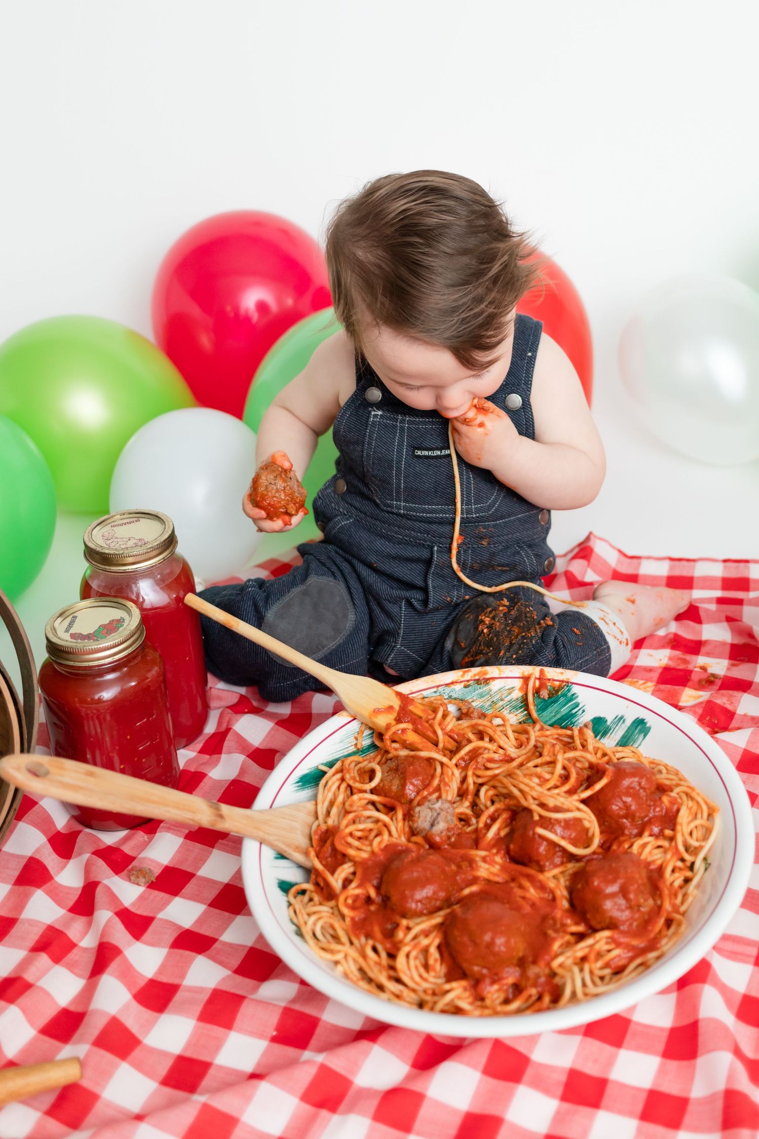 Spaghetti and meatballs dinner setup with colorful balloons and mason jars on a checkered picnic blanket.