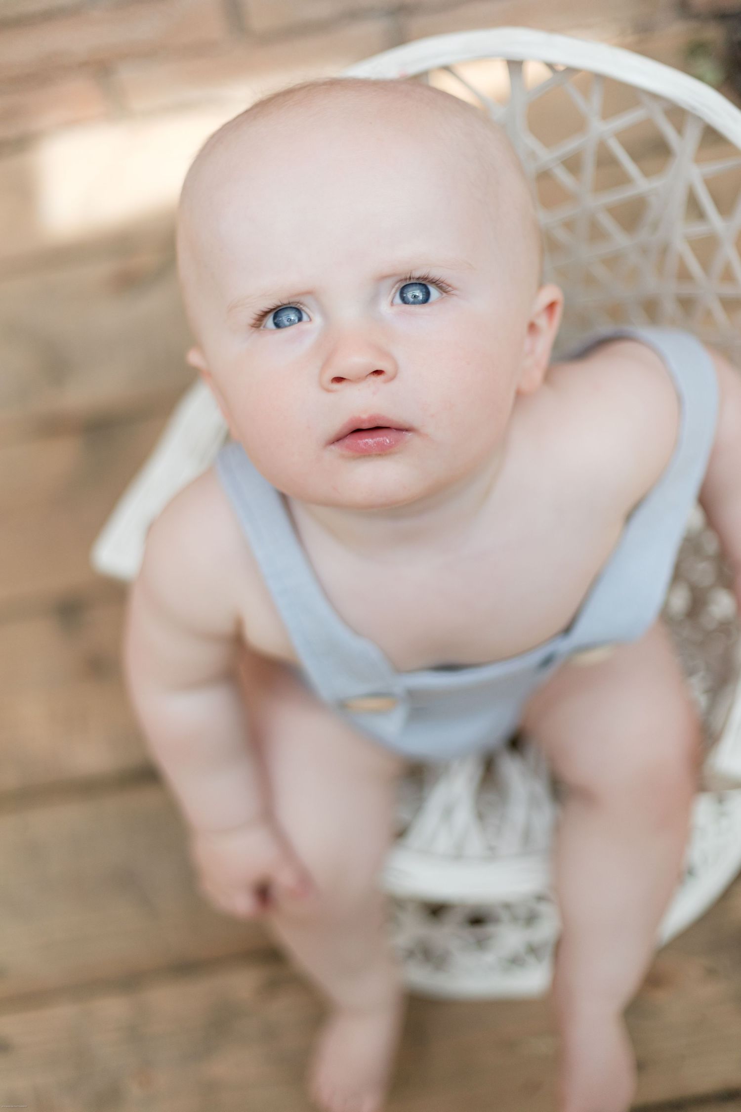 Baby wearing light blue romper sits in white wicker chair against brick wall.