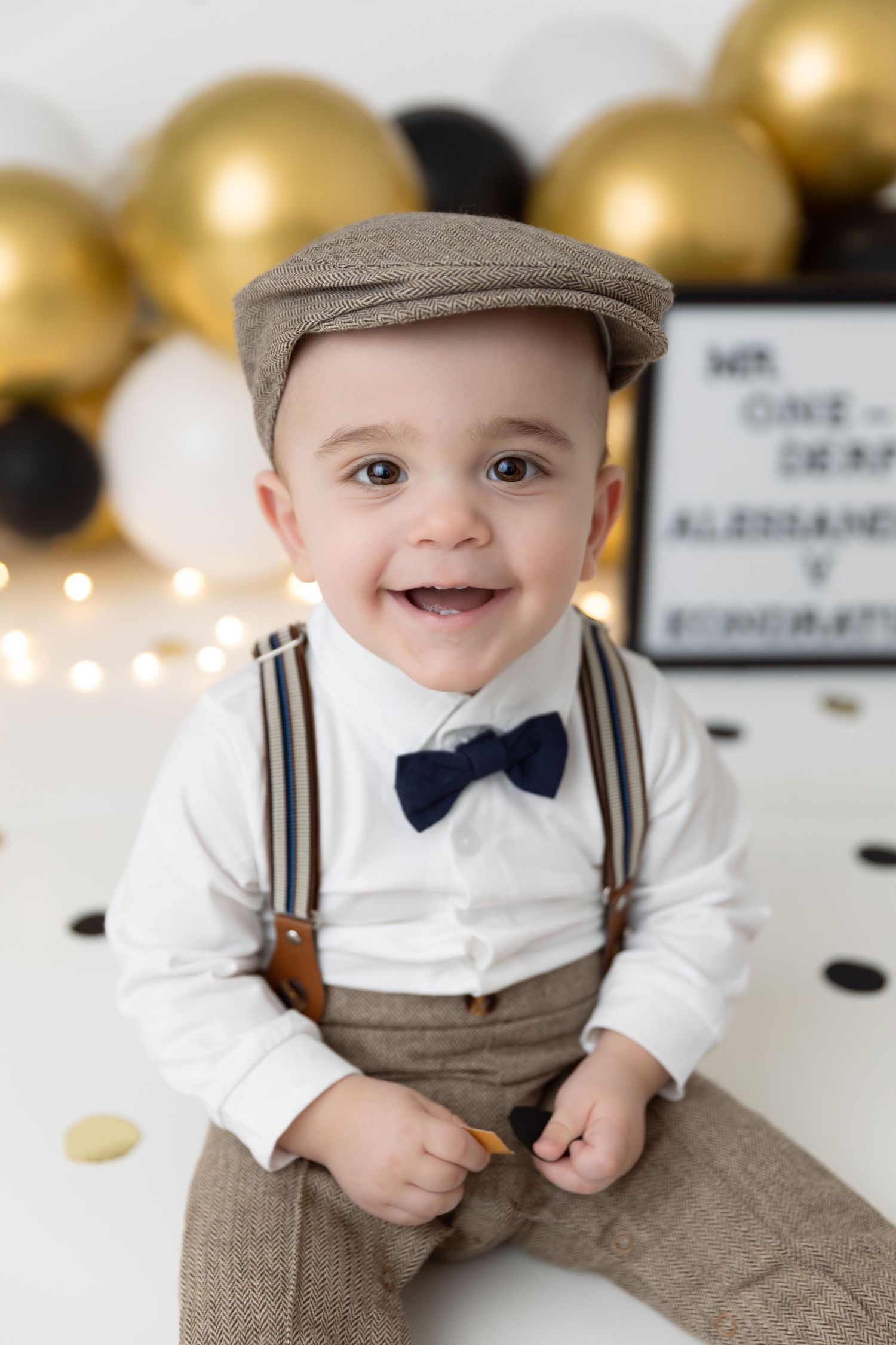 Adorable young child dressed in vintage style outfit with suspenders, bow tie and newsboy cap against gold balloon backdrop.