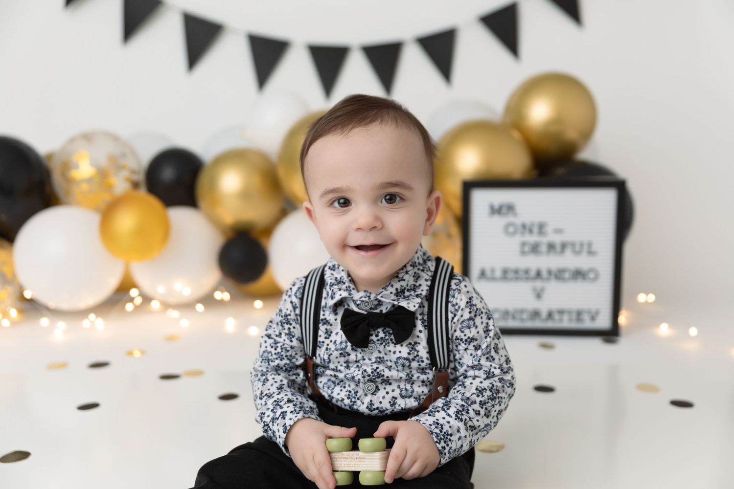 Baby in black and white patterned outfit sits on floor with gold and white balloons and party decorations in background.