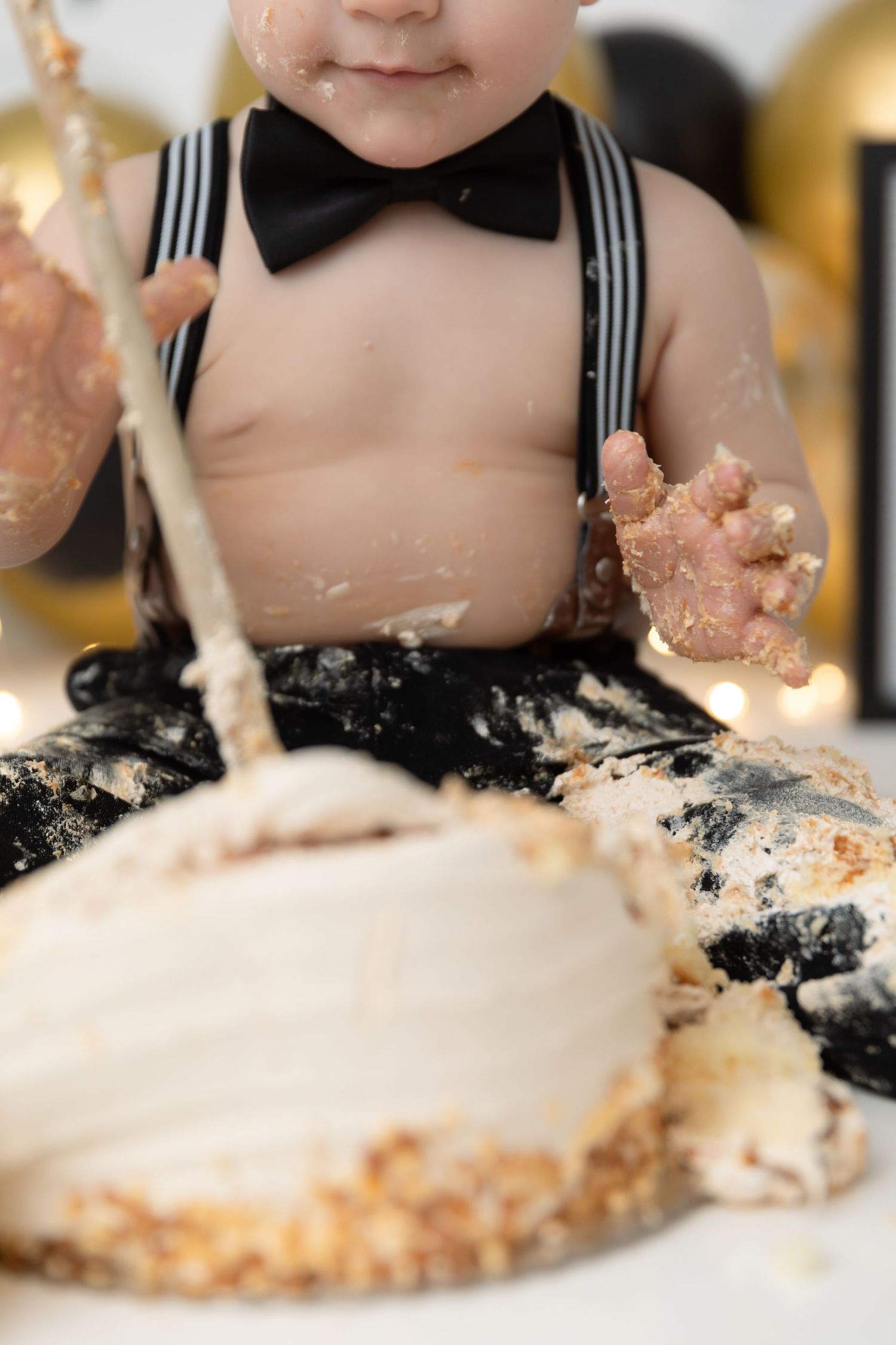 Close-up view of a messy cake smash photo featuring suspenders and bow tie during a first birthday celebration.