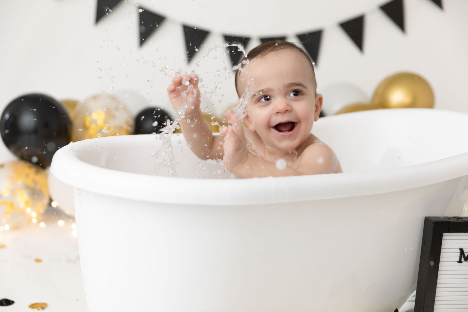 Happy baby splashing in white bathtub with black and gold balloons and party decorations in background.