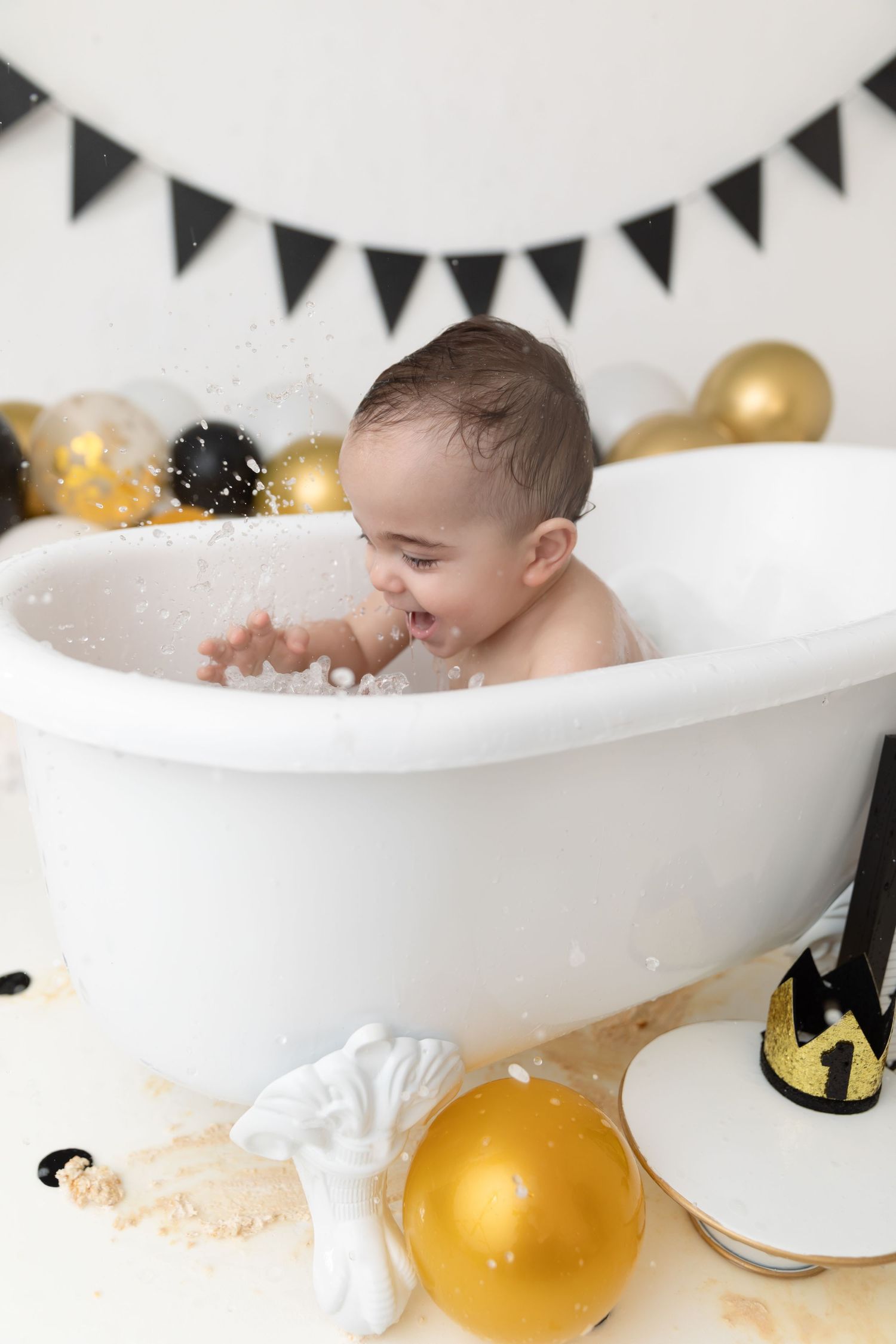 Laughing baby splashes in white bathtub surrounded by gold balloons and black party decorations against white backdrop.