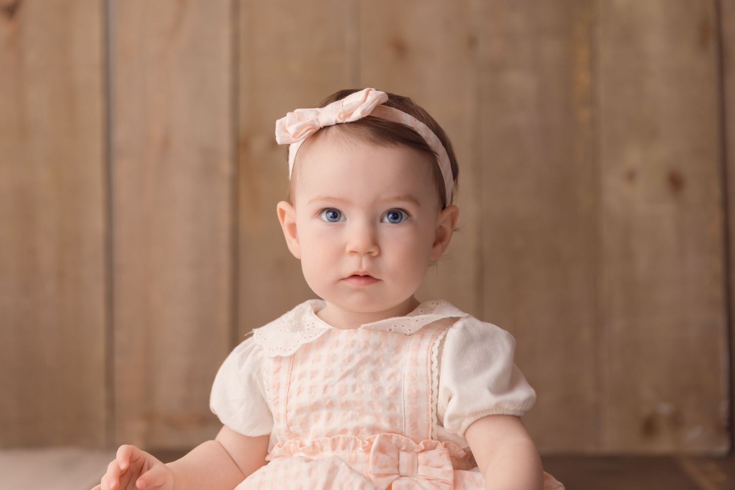 Small child wearing a peach-colored dress with white puff sleeves and matching headband against a wooden backdrop.