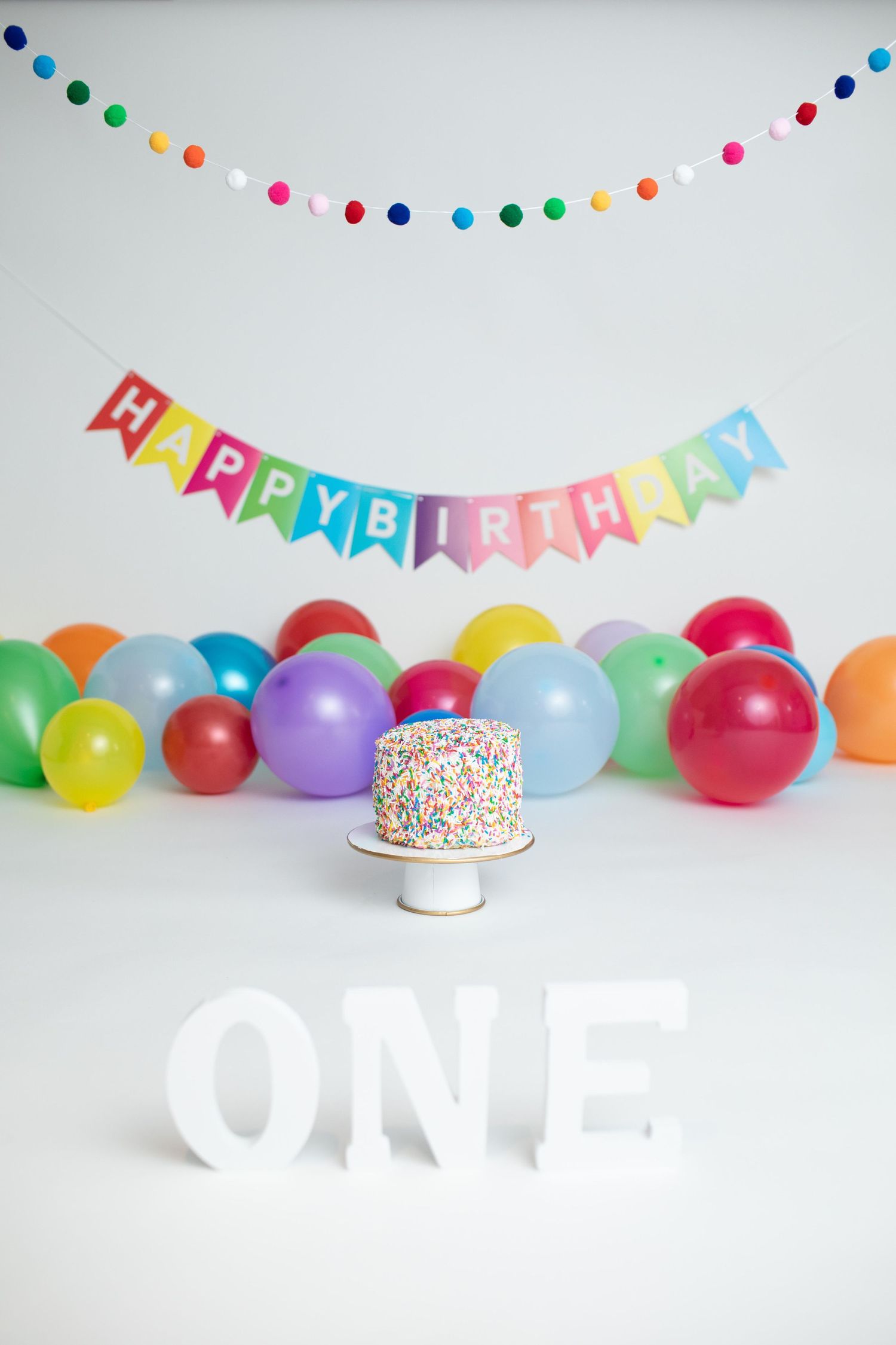 Colorful first birthday party setup with rainbow balloons, sprinkle cake and 'ONE' letters.