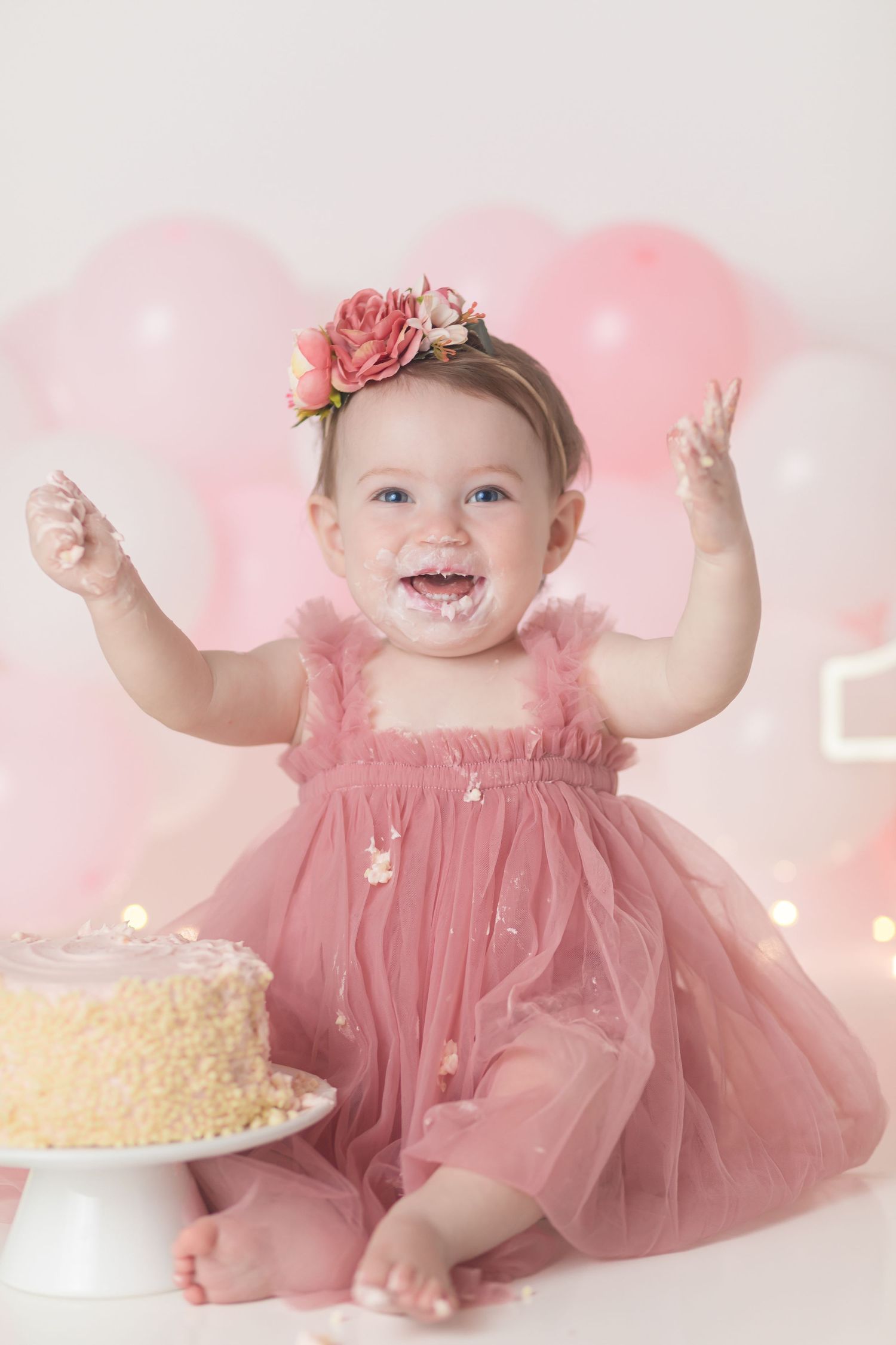 Baby in pink tulle dress celebrates with first birthday cake against pink balloon backdrop.