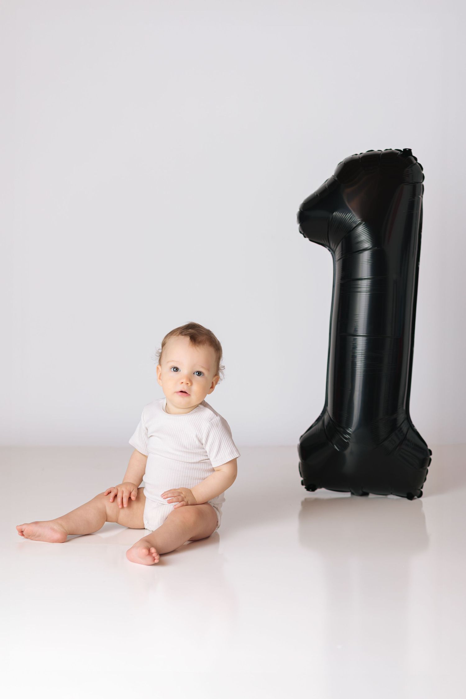 A baby in white onesie sits next to large black number one balloon on white background.