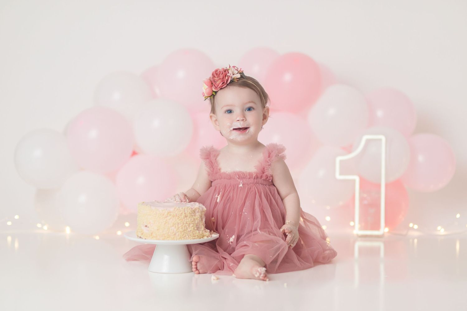 First birthday photoshoot setup with pink balloons, glowing lights, yellow cake on stand, and floral headband decor.