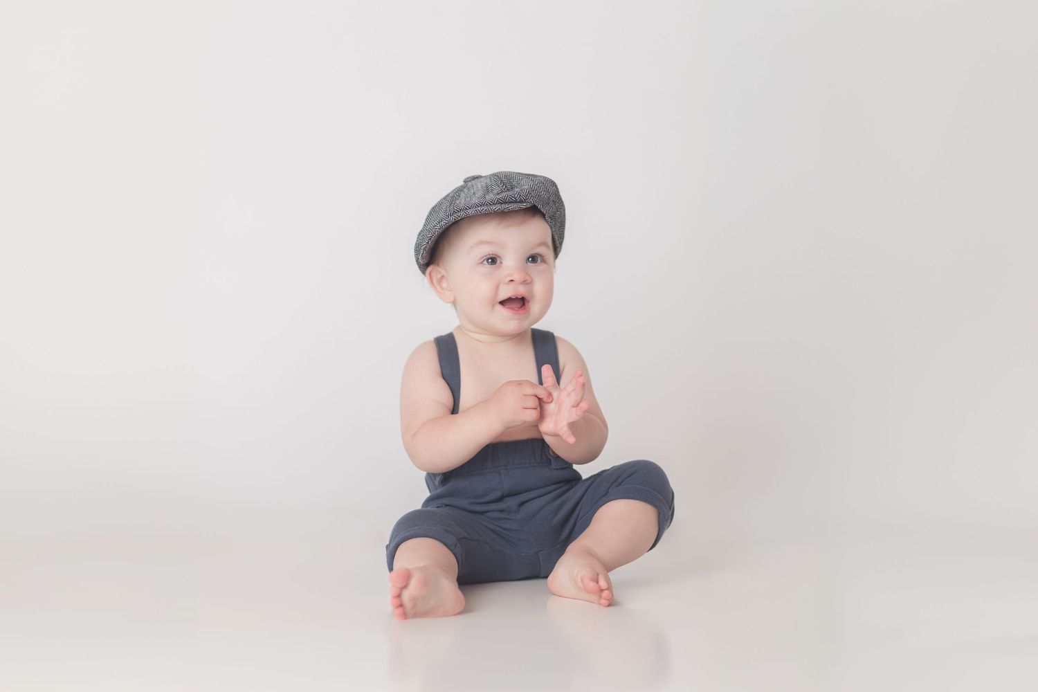 Small baby wearing a gray cap and dark overalls sits on a white studio backdrop with a cheerful expression.