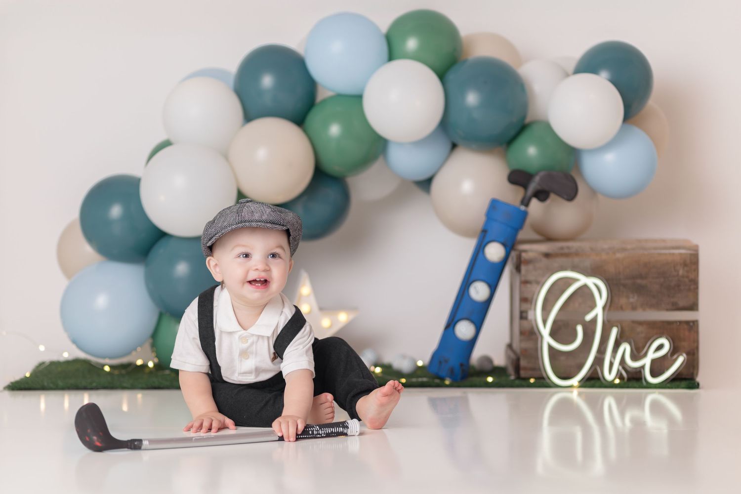 Baby in formal wear sits near a balloon arch and wooden 'One' sign for first birthday photoshoot.