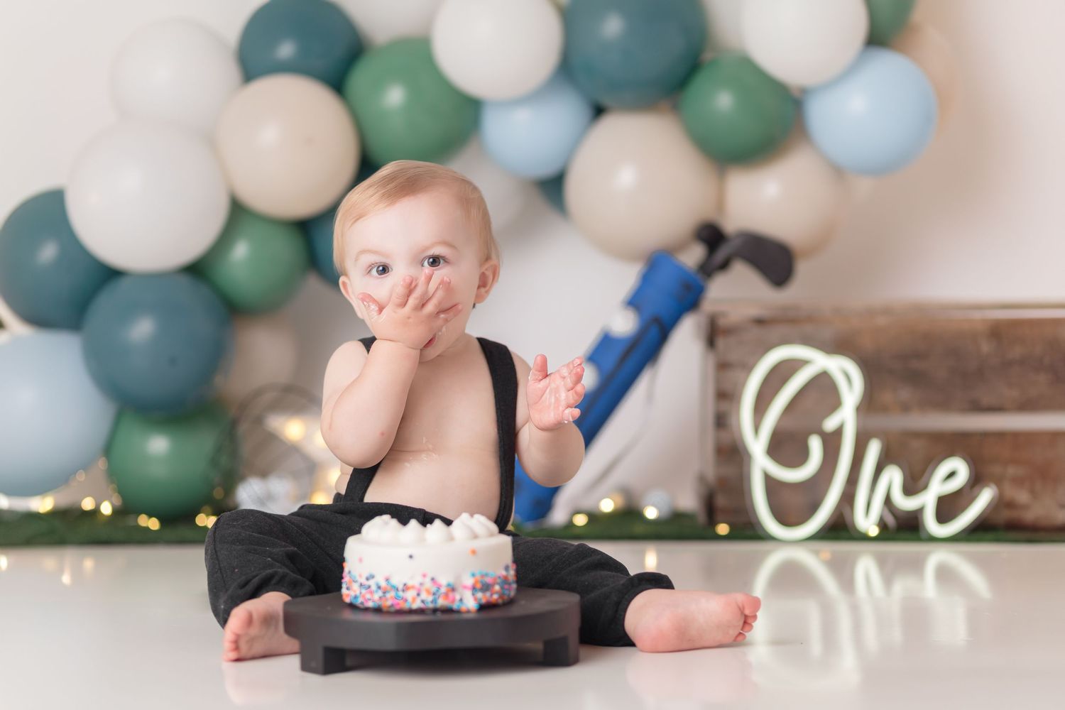 First birthday cake smash photo with blue and white balloon arch backdrop and lit 'One' sign.