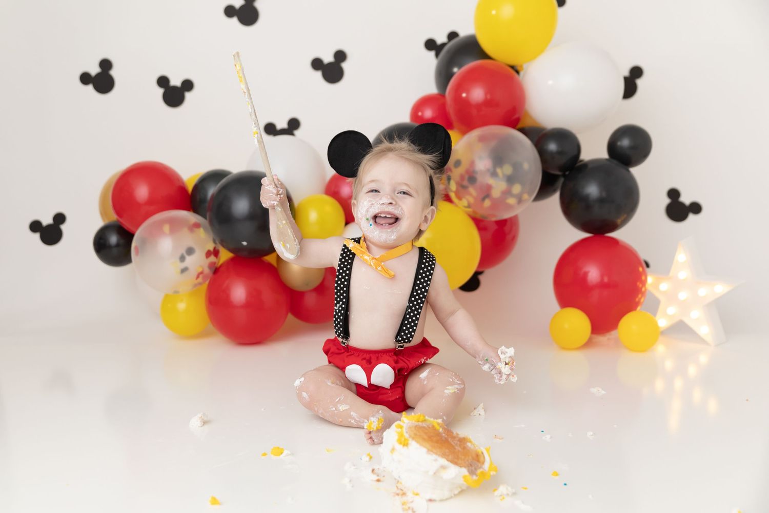 Mickey Mouse themed first birthday cake smash photo with red, yellow and black balloons and Mickey ears on white backdrop.