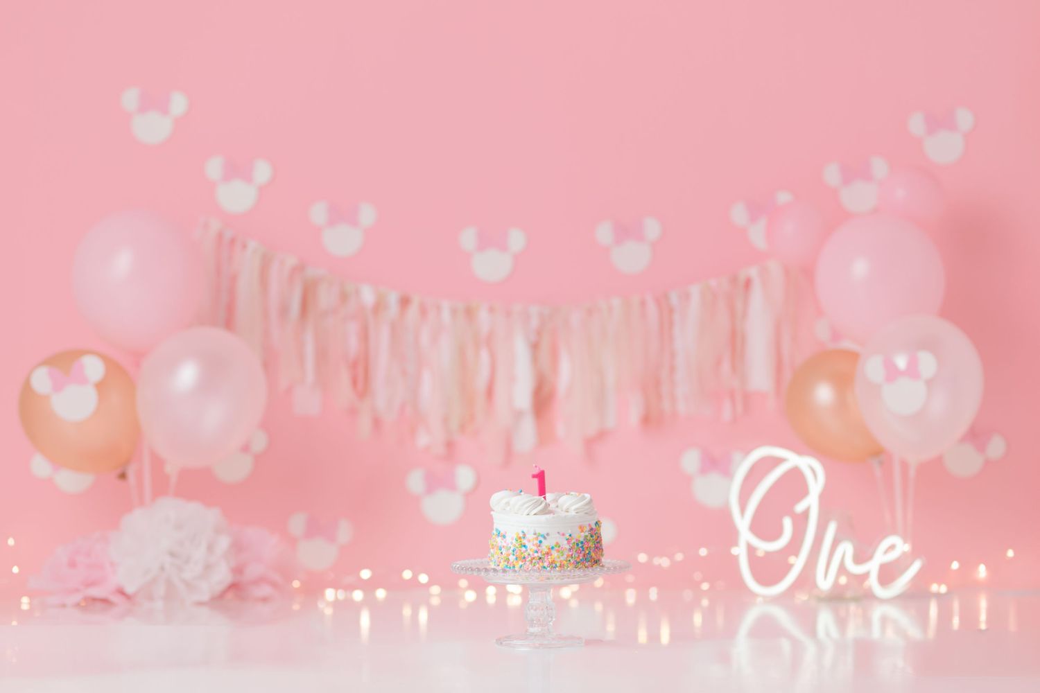Pink first birthday party setup with balloons, banners and cake on a pink background.
