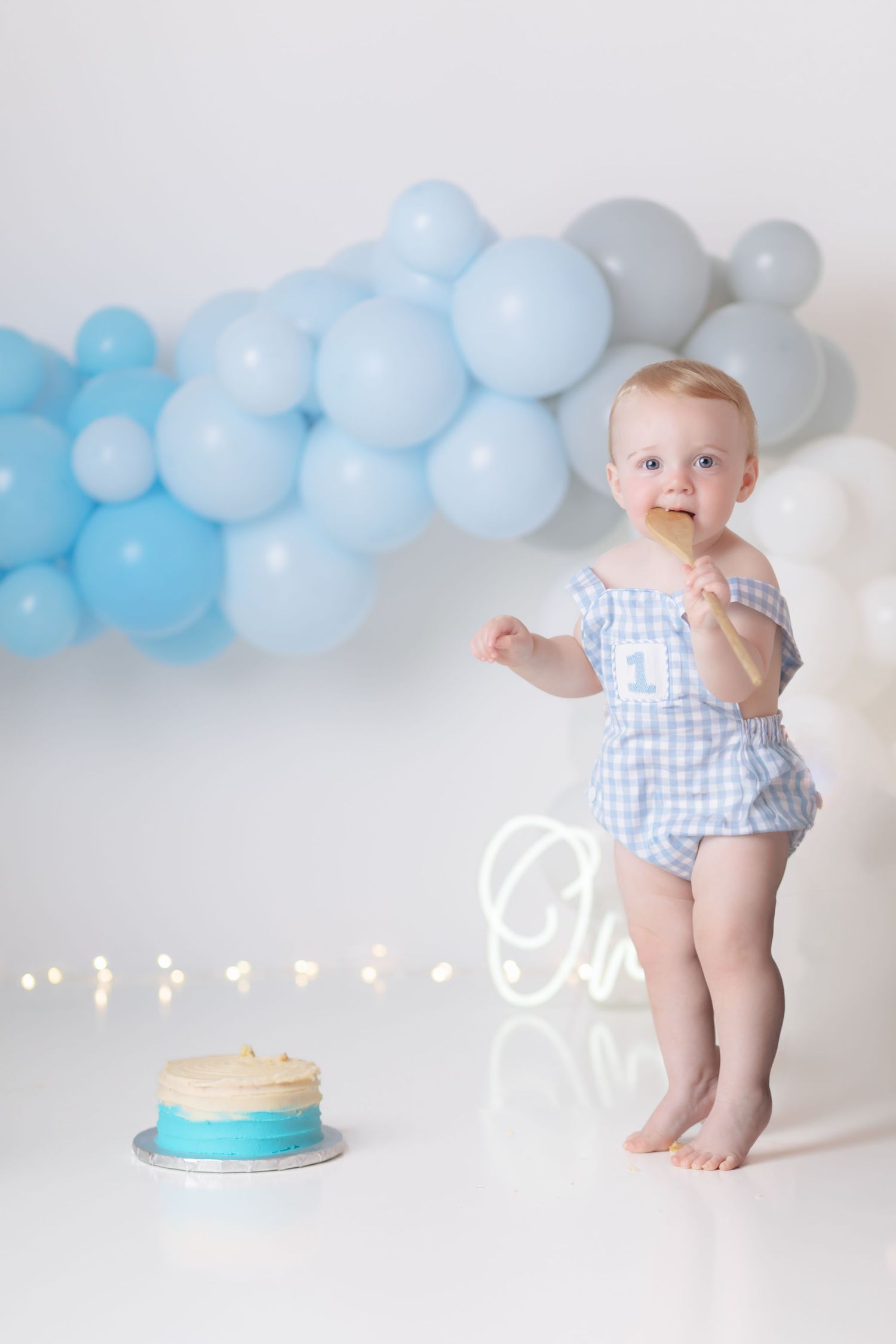 Baby in blue gingham romper stands near cake with blue balloon arch decoration.